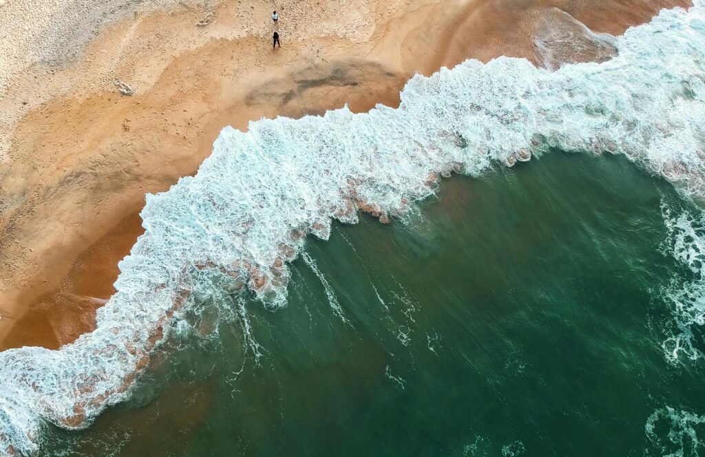 pexels-photo-982263-982263 Aerial shot of ocean waves crashing onto a sandy beach with two people in the distance.